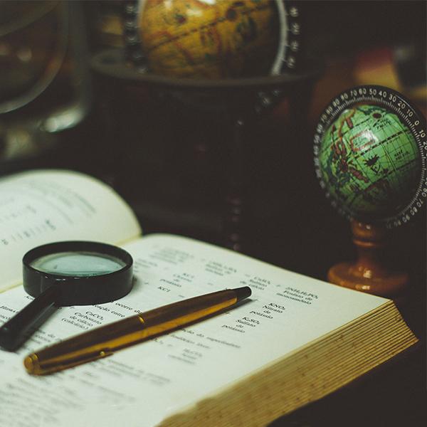 Photo of a magnifying glass and a pen atop a textbook. A globe is in the background